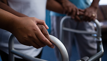 A close-up of a person's hand gripping a walker with another person assisting in the background.