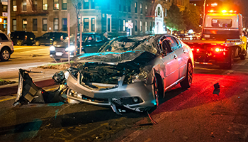 A damaged car at a nighttime accident scene with emergency vehicle lights in the background.